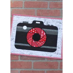 Nahaufnahme des zweiten Kamera-Mini-Quilts mit rot-weißem Blendenmuster, Landkarten-Hintergrundstoff und rot-weißem Zackenrand-Binding vor einer Backsteinmauer.